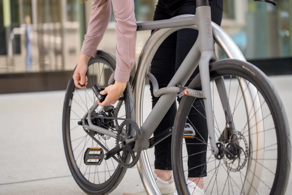 Frau in Anzughose und Hemd schließt Fahrrad mit tex–lock eyelet in electric grey als Leasing-Schloss an Fahrradstander ab.